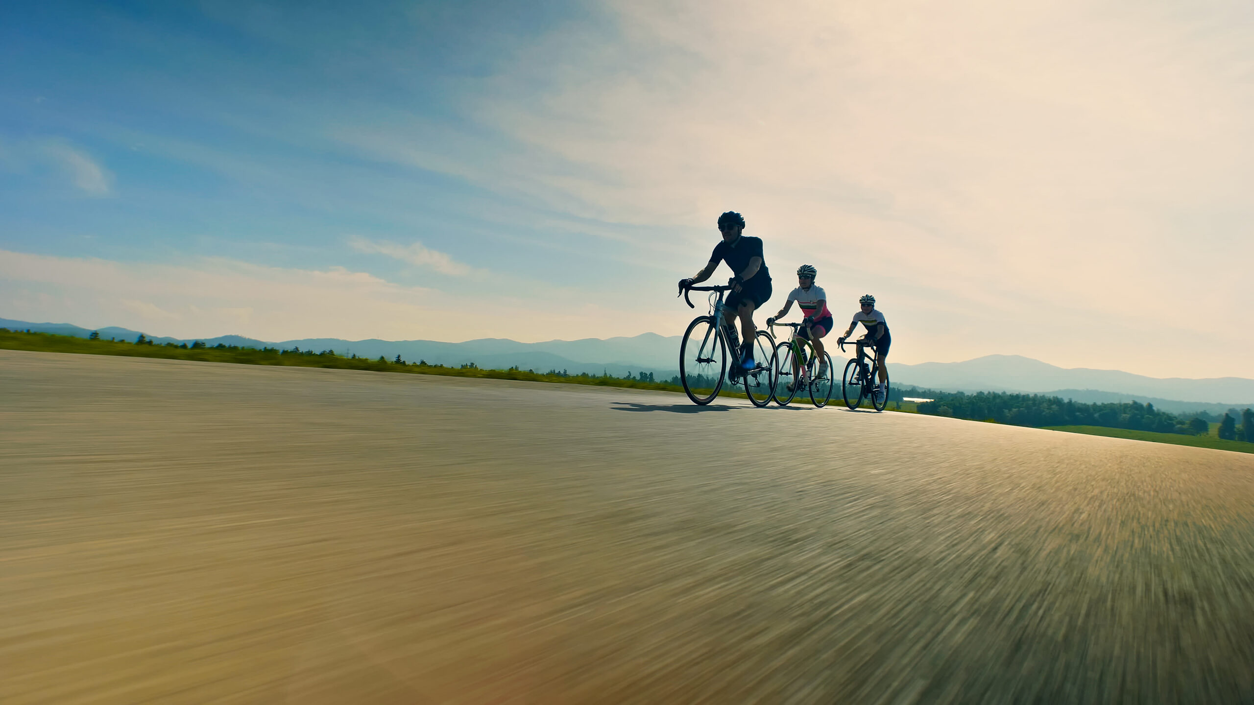 Three people pedaling bicycles are leading a road cycling race through a valley with tree-covered hillsides in the distance. The image also shows the forward motion and a blur from the speed at which the cyclists are traveling. The image symbolizes PE-backed leadership requiring a different approach and pace.